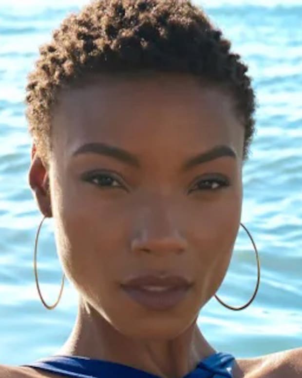 Iyonna Fairbanks poses in front of the ocean in a deep blue bikini and oversized hoop earrings.